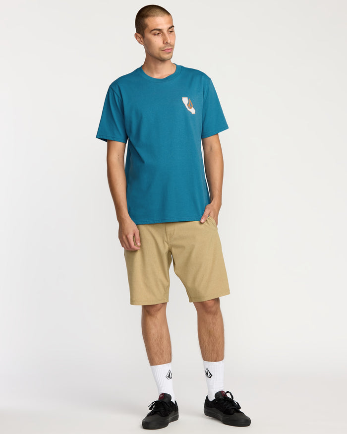 A man with short hair stands against a plain background wearing the Volcom Men's California Dreaming Short Sleeve Tee in teal with a soft-hand chest print, tan shorts, white socks, black sneakers, and his hands in his pockets.