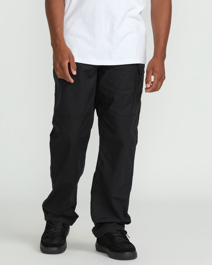 A person wears Volcom Men's Caliper II Relaxed Work Pants in black, paired with a white T-shirt and black sneakers, standing against a plain light background. Only the lower half of their body is visible.
