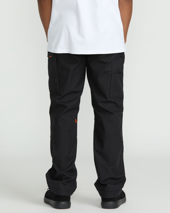 A person wearing a white T-shirt and Volcom Men's Caliper II Relaxed Work Pants in black stretch canvas with cargo pockets and small orange logo details stands facing away from the camera, paired with black shoes.