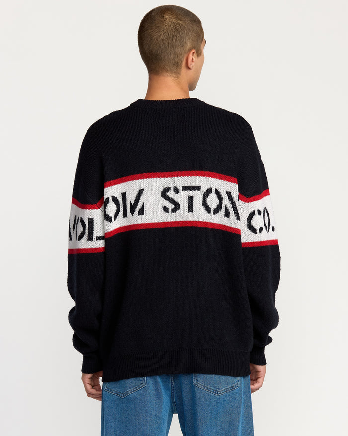 A person with short hair, seen from behind, wears the Volcom Men's Carwash Sweater—black with a white and red horizontal stripe featuring lettering—paired with blue jeans.