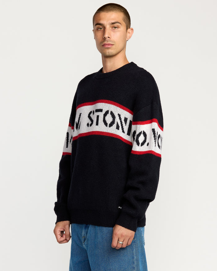 A person with short hair wears the Volcom Men's Carwash Sweater—black with red and white stripes and bold lettering—paired with blue jeans, standing against a plain white background.