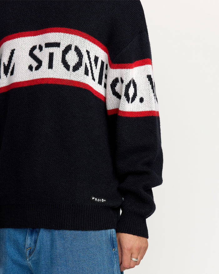 A person is shown wearing the Volcom Men's Carwash Sweater, which features black fabric with red and white stripes and bold text. The sweater is paired with blue jeans, displaying only the torso and one hand.