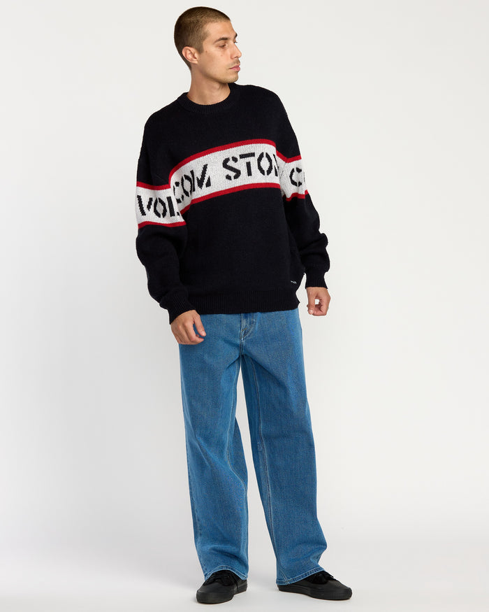 A person stands against a plain white background, looking to the side, wearing the Volcom Men's Carwash Sweater with VOLCOM STONE text, red and white stripes, blue wide-leg jeans, and black shoes.