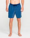 A person standing barefoot, wearing Volcom Men's Center Elastic Waist Trunks in blue with a black drawstring—classic men's swimwear—shown from shoulders to feet against a plain light background.