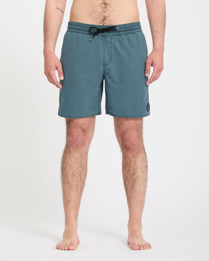 A man stands barefoot against a white background, wearing Volcom Men's Center Trunks 17 teal swim shorts that reach above the knee. His arms rest at his sides and the upper part of his body is partially out of frame.