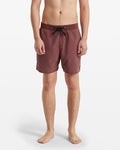 A person stands barefoot, facing forward against a white background, wearing Volcom Men's Center Elastic Waist Trunks in maroon with a black drawstring, elastic waist, and a small patch on the left leg.