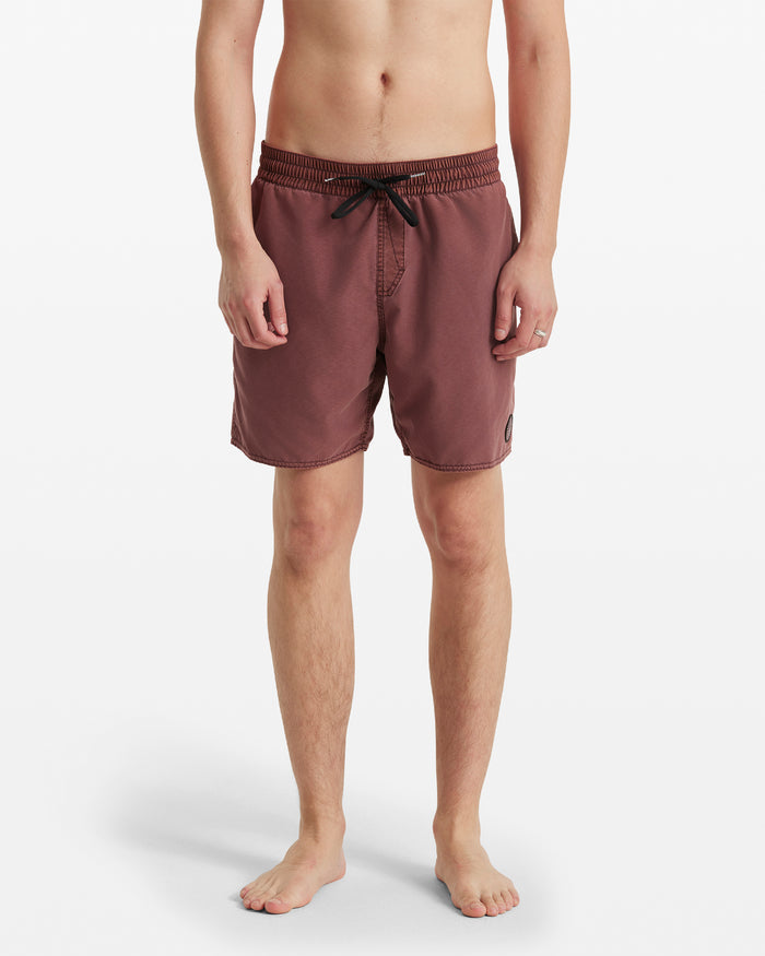 A person stands barefoot, facing forward against a white background, wearing Volcom Men's Center Elastic Waist Trunks in maroon with a black drawstring, elastic waist, and a small patch on the left leg.