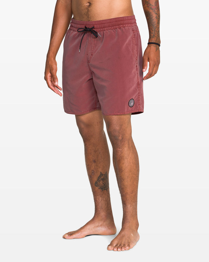 A man stands barefoot against a white background in Volcom Men's Center Elastic Waist 17" Trunk, faded red with a black drawstring and circular logo on the left leg. Made from recycled polyester, he shows tattoos on his left arm and leg.