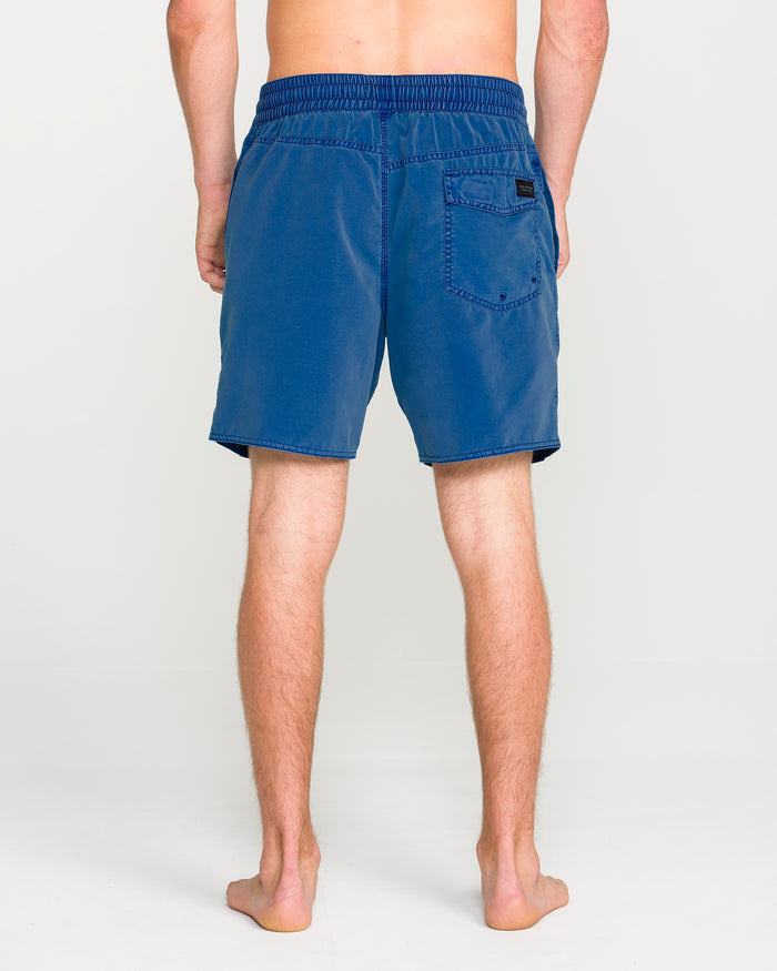 A person stands barefoot facing away, wearing blue Volcom Men's Center Elastic Waist Trunks featuring an elastic waistband and a back pocket. The background is plain light gray.
