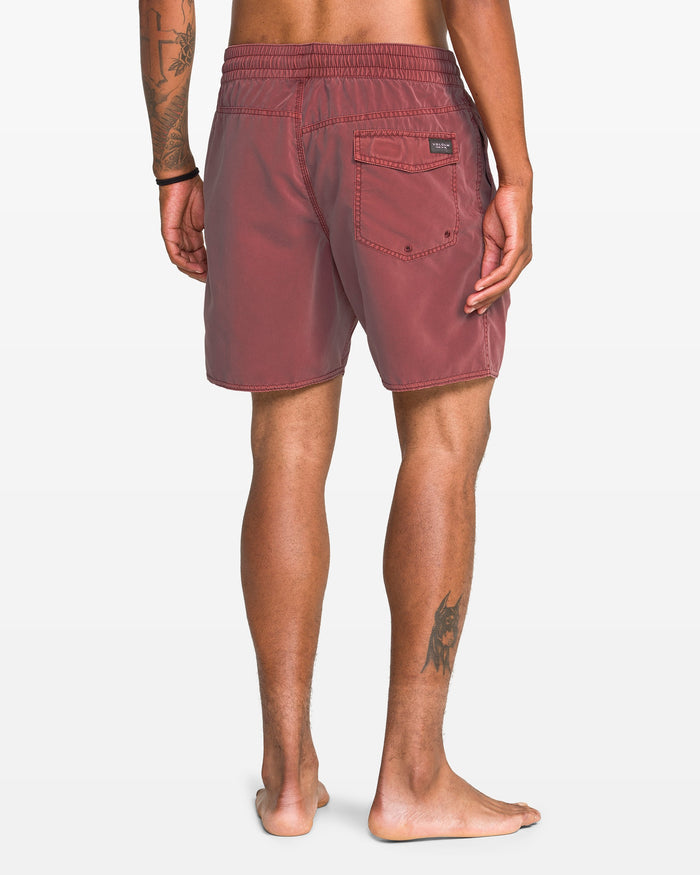 A barefoot person faces away in Volcom Men's Center Elastic Waist 17" Trunk—red, recycled polyester shorts with a back pocket. Tattoos on their left arm and right calf are visible against a plain white background.