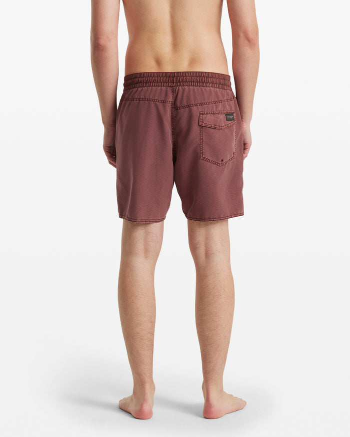 A person stands barefoot, facing away, wearing Volcom Men's Center Elastic Waist Trunks in maroon polyester blend with an elastic waist and back pocket. The mid-length trunks stand out against the plain white background.