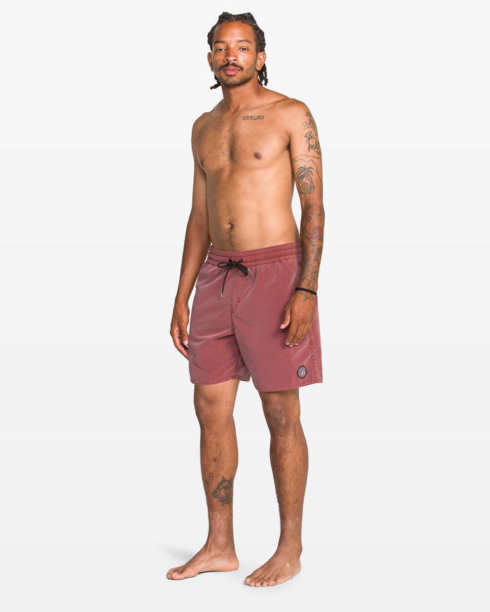 A man with braids, tattoos, and facial hair stands barefoot against a white background wearing Volcom Men's Center Elastic Waist 17" Trunk in maroon—featuring recycled polyester, mechanical stretch, a black drawstring, and a logo on the left leg.