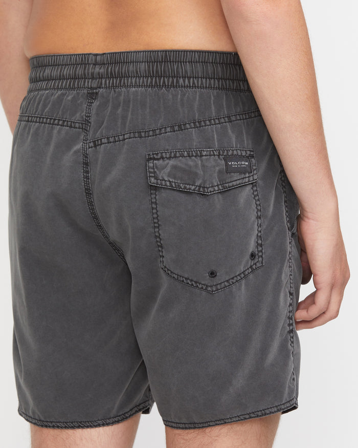 A person wears Volcom Men's Center Elastic Waist 17" Trunk swim shorts in dark gray, made from recycled polyester with an elastic waistband and back pocket, standing against a plain white background.