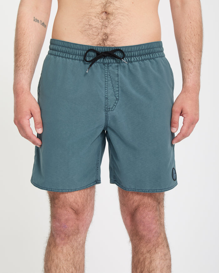 A person stands facing forward in Volcom Men's Center Trunks 17, teal swim shorts with side pockets and a small patch logo on the front left leg. Their arms rest at their sides against a plain white background.