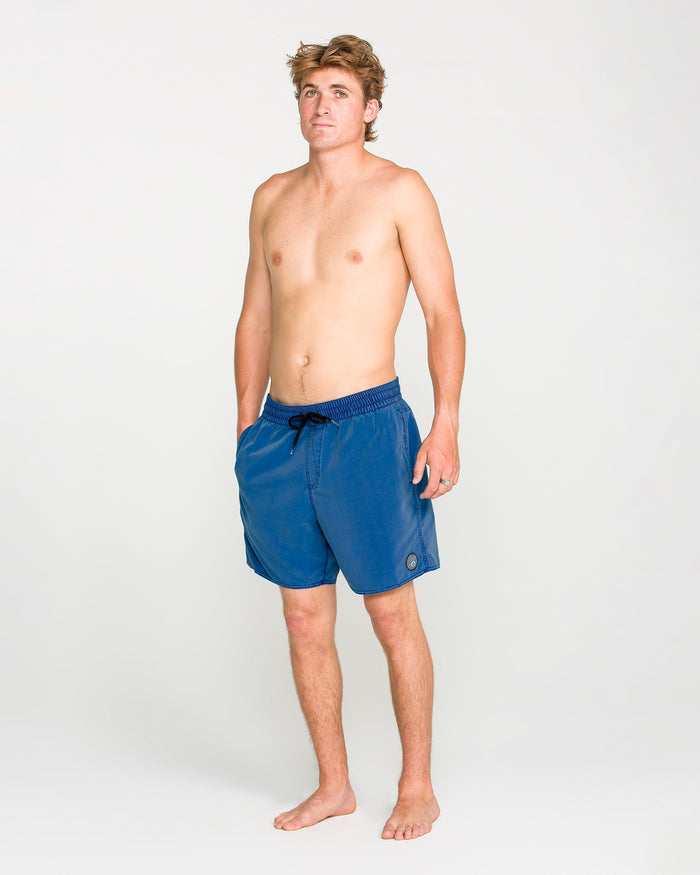 A barefoot man with light brown hair stands against a plain background, wearing Volcom Men's Center Elastic Waist Trunks in blue and no shirt. His hands are in his pockets, and he is looking slightly upward and to the left.