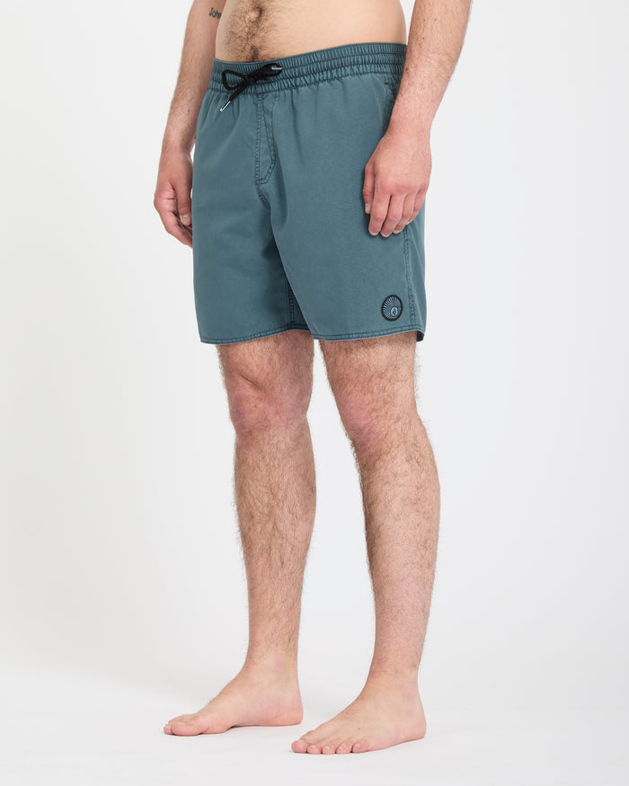 A man stands barefoot on a white background, wearing Volcom Men's Center Trunks 17 in teal, featuring a black drawstring and a small round logo near the left leg hem. His upper body is not visible.