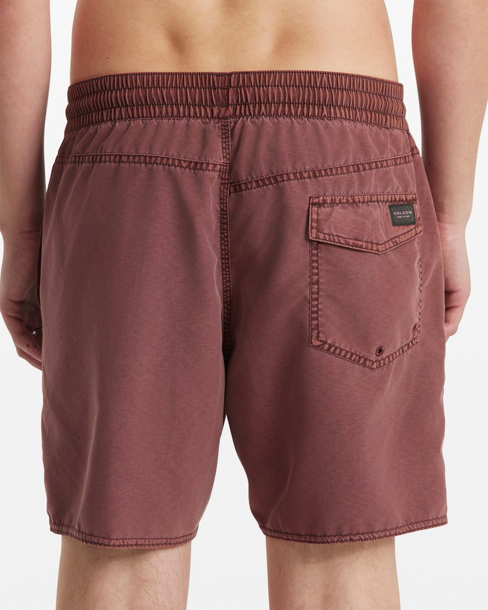 A person shown from the waist down, viewed from behind, wears maroon Volcom Men's Center Elastic Waist Trunks with mid-length cut and a single back pocket.
