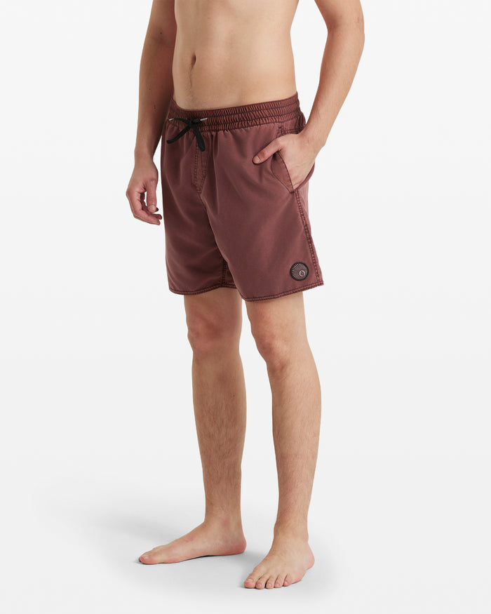 A person stands barefoot against a white background wearing Volcom Men's Center Elastic Waist Trunks in mauve, featuring a mid-length cut, side pockets, and a comfortable polyester blend.