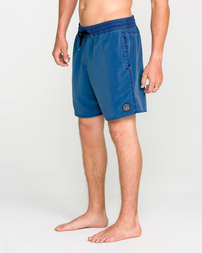 A person stands barefoot on a light background, wearing Volcom Men's Center Elastic Waist Trunks in blue with a 17-inch length, featuring a black drawstring and a circular patch on the left leg. The upper body is not visible, drawing attention to the stylish swimwear.