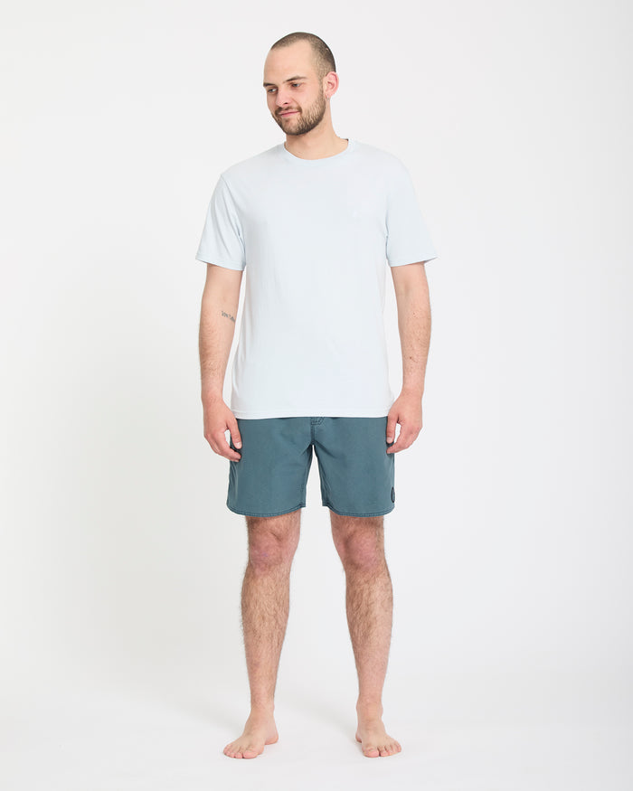 A man with a short beard stands barefoot against a white background, wearing Volcom Men's Center Trunks 17 and a light blue t-shirt, looking slightly to the side with a relaxed expression.