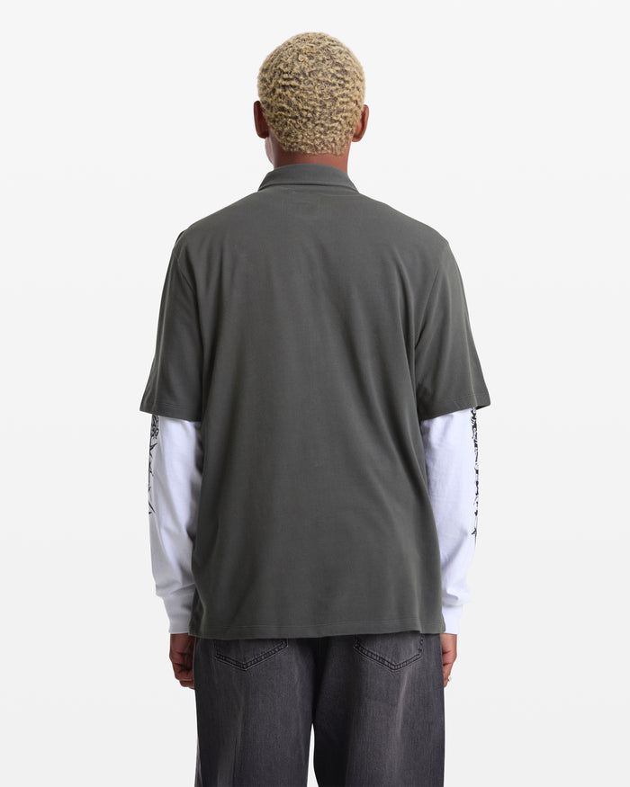 A person with short, curly blond hair is seen from behind wearing the Volcom Men's Chavo Button Down Short Sleeve shirt in dark gray textured cotton pique over a white long-sleeve shirt and dark gray jeans, against a plain white background.