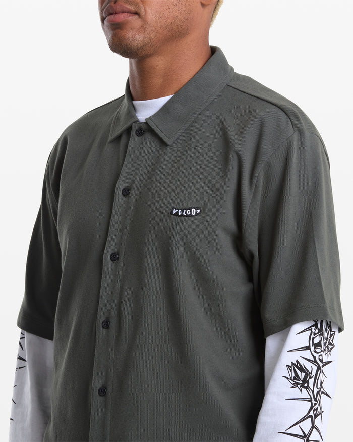 A person wears the Volcom Men's Chavo Button Down Short Sleeve shirt in dark green cotton pique, featuring a logo patch, layered over a white long-sleeve with black graphic artwork on the sleeves.