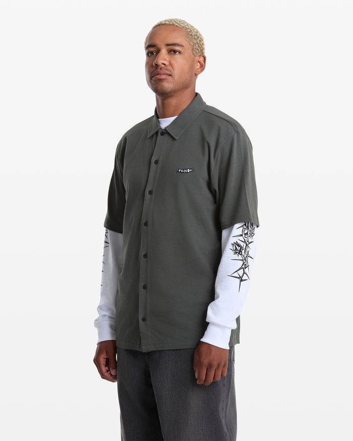 A person with short, bleached hair wears a dark green Volcom Men's Chavo Button Down Short Sleeve shirt over a white long-sleeve with black sleeve designs and dark pants, standing against a plain white background.