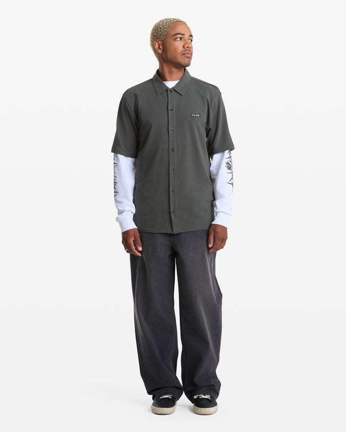 A person with short blonde hair wears a Volcom Men's Chavo Button Down Short Sleeve over a long-sleeve white printed top, loose dark jeans, and black sneakers, standing against a plain white background.