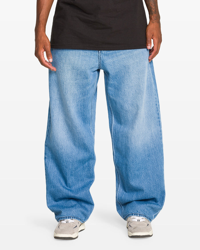 A person wears Volcom Men's Chillow Denim Vs jeans, paired with white sneakers and a black t-shirt, showing only their torso and legs against a plain white background.
