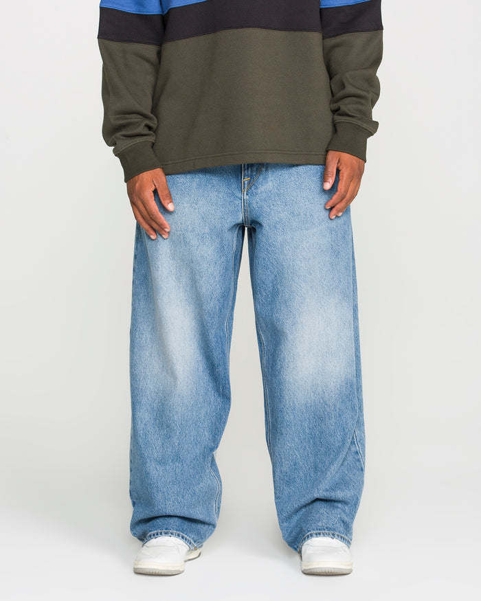 Model showcases the Volcom Men's Chillow Denim Pants in Euro Light Blue—a baggy, low-rise fit—paired with a long-sleeve shirt and white shoes. Only the body from shoulders down is visible, photographed against a plain white background.