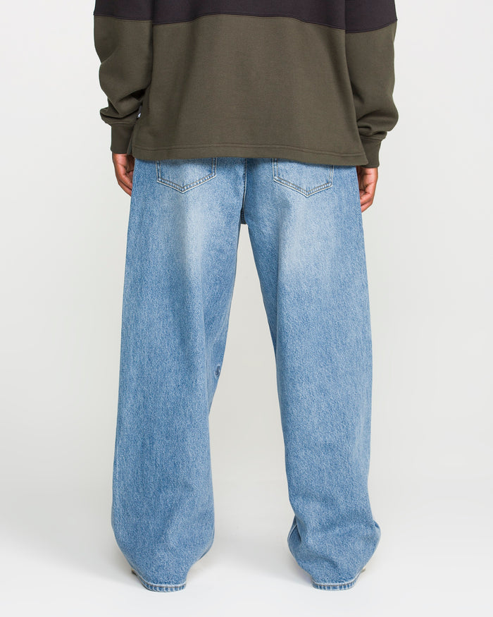 A person stands with their back to the camera against a plain white background, wearing a dark olive green sweatshirt and Volcom Men's Chillow Denim Pants in a loose-fitting, Euro Light Blue wash.