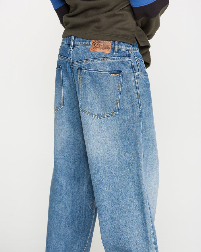 A person wearing Volcom Men's Chillow Denim Pants, featuring a relaxed fit in blue denim with visible back pockets and a brown patch on the waistband, is photographed from behind against a plain background.