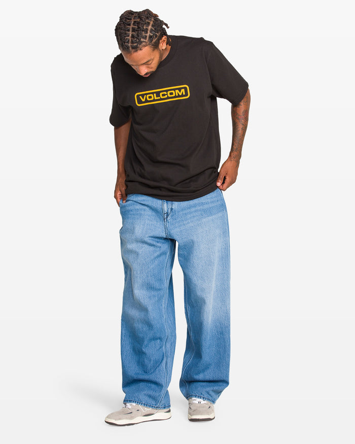 A person wears a black Volcom t-shirt, Men’s Chillow Denim Vs jeans by Volcom, and white sneakers, standing with hands in pockets against a plain white background.