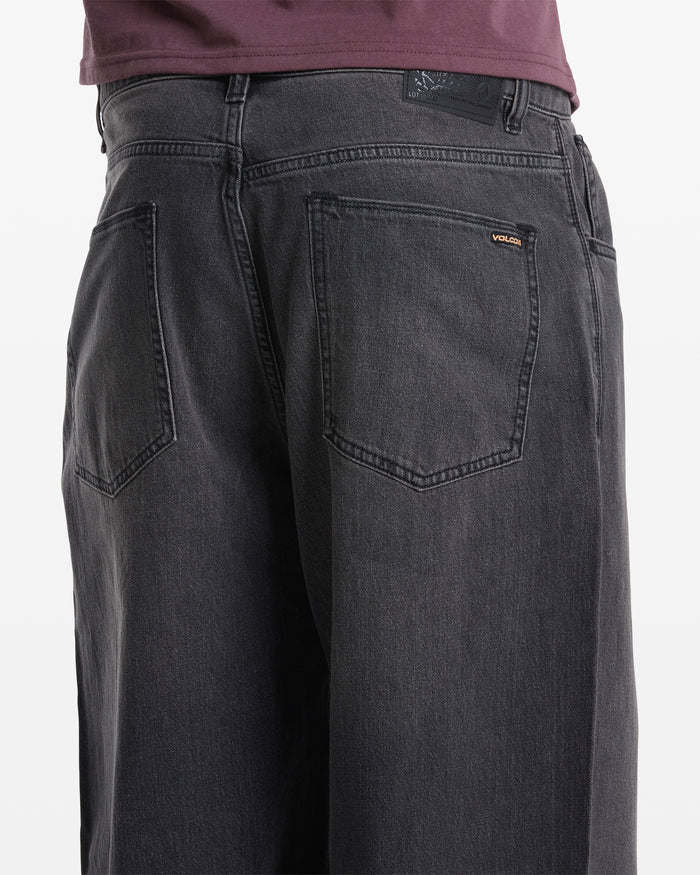 Close-up of the back of a person wearing Volcom Men's Chillow Denim in dark gray, featuring a relaxed fit, two back pockets, and a small Volcom logo tag above the right pocket, paired with a dark purple shirt.