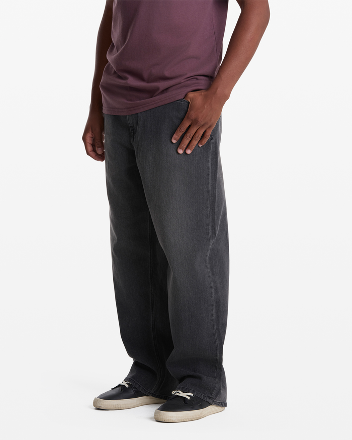 A person wearing Volcom Men's Chillow Denim—loose-fitting dark gray jeans—stands against a white background, paired with a mauve t-shirt and black-and-white sneakers. Their head is not visible in the image.