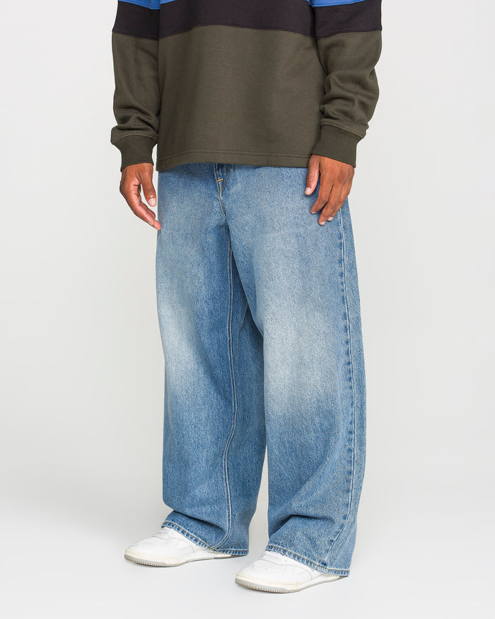 A person wearing a dark sweater, Volcom Men's Chillow Denim Pants in Euro Light Blue, and white sneakers stands against a plain background. Only the lower half of the body is visible.