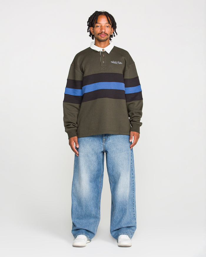 A man stands facing forward, wearing a dark green rugby shirt with blue and black horizontal stripes, Volcom Men's Chillow Denim Pants, and white sneakers against a plain background.