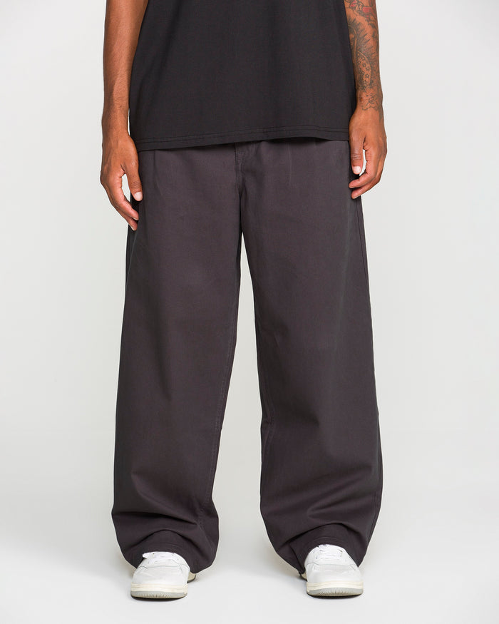 A person, shown from the waist down against a plain white background, is wearing Volcom Men's Chillow Pleat Elastic Waist Pants in a loose dark gray fit, paired with a black shirt and white sneakers.