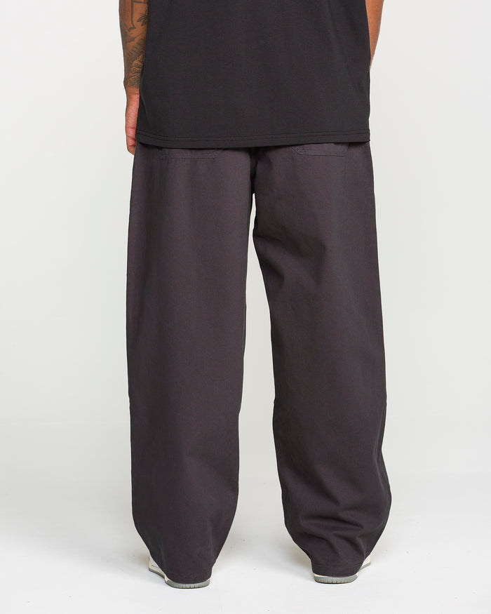 A person stands facing away against a plain white background, with only their lower body visible, wearing Volcom Men's Chillow Pleat Elastic Waist Pants in black cotton twill with a wide-leg silhouette and a black shirt.