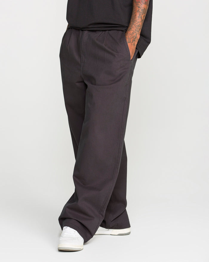 A person wearing Volcom Men's Chillow Pleat Elastic Waist Pants, paired with white sneakers and a black shirt, stands with hands in pockets. Only the lower torso and legs are visible against a plain background.
