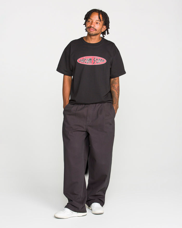 A man stands against a plain background wearing a black Volcom Stone t-shirt and Volcom Men's Chillow Pleat Elastic Waist Pants, paired with white sneakers, his hands in his pockets and a calm expression on his face.