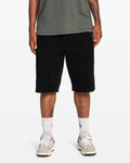 A person stands against a plain white background, showcasing their lower body in Volcom Men's Chillow 24" Shorts (antique black), paired with a green t-shirt, white socks, and white sneakers.