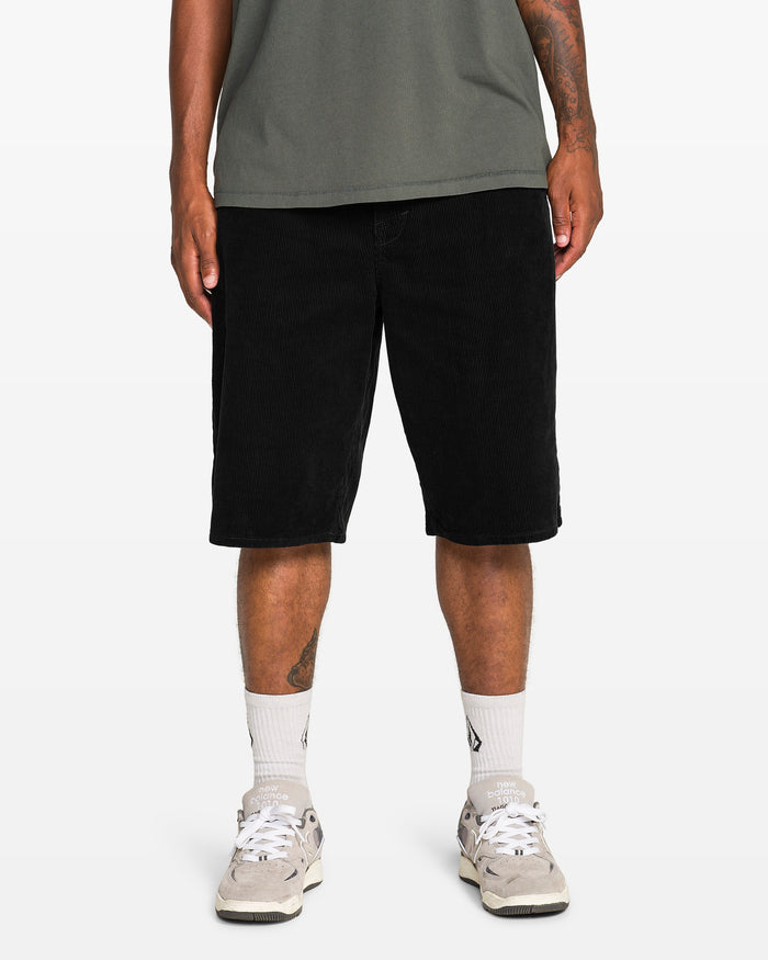 A person stands against a plain white background, showcasing their lower body in Volcom Men's Chillow 24" Shorts (antique black), paired with a green t-shirt, white socks, and white sneakers.