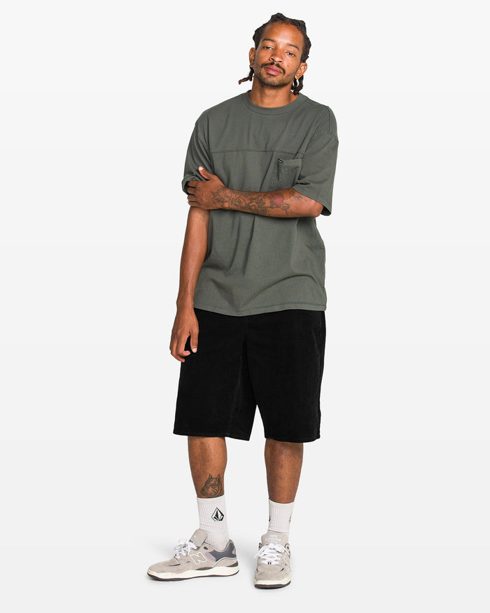 A man with braided hair stands against a white background, wearing a loose dark green tee, Volcom Men's Chillow 24" Shorts in antique black, white socks, and gray sneakers. He has one arm crossed and looks relaxed.