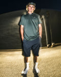 Wearing the Volcom Men’s Chillstone Short Sleeve Polo in green, along with navy shorts, a cap, white socks, and sneakers, a person stands outside at night against a blurred background with a lighting effect.