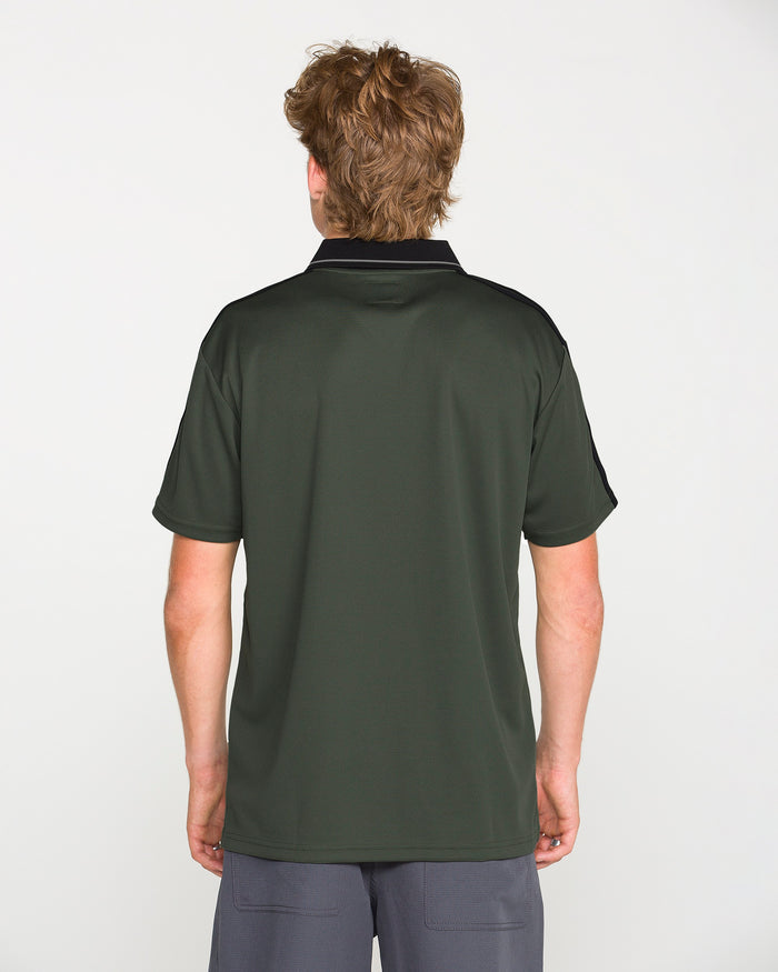 A person with light brown hair stands facing away, wearing the Volcom Men's Chillstone Short Sleeve Polo in dark green with a black collar and shoulders featuring subtle yarn-dye stripes, paired with gray shorts, against a plain white background.