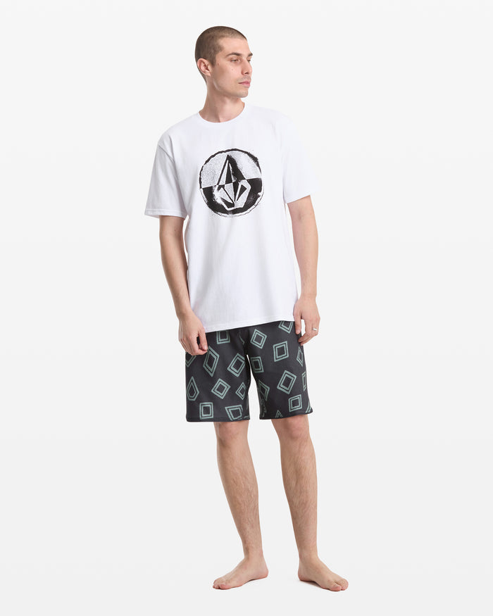 A man stands barefoot in a Volcom Men's Circle Split Short Sleeve Tee with a bold black logo on the front and black shorts with a blue square pattern, looking slightly left against a plain white background.