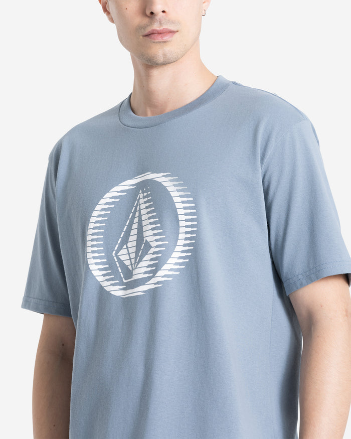 A person wearing a Volcom Men's Circle Stone Short Sleeve Tee in light blue, made from 100% cotton, featuring a white geometric logo with circular lines on the chest. The photo shows their upper body against a plain light background.
