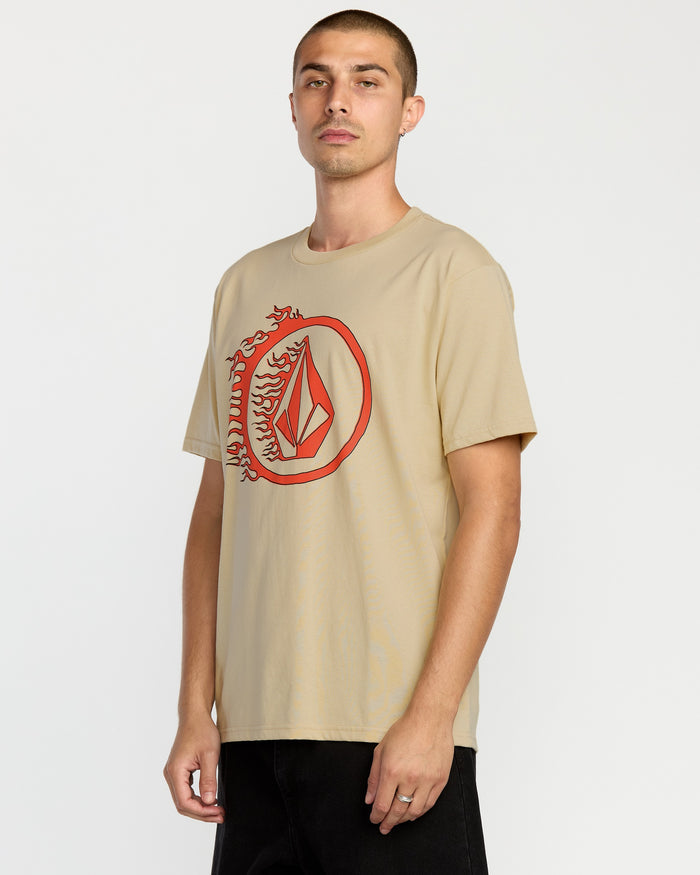 A man with short hair wears the Volcom Men's Circle Stone Short Sleeve Tee in beige, featuring a large red geometric logo with flame details on the left chest, made from premium ring-spun cotton, standing against a plain white background.