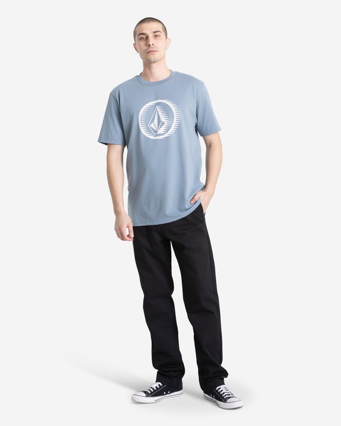 A person wearing a Volcom Men's Circle Stone Short Sleeve Tee, black pants, and black Converse sneakers stands with one hand in their pocket against a plain white background.